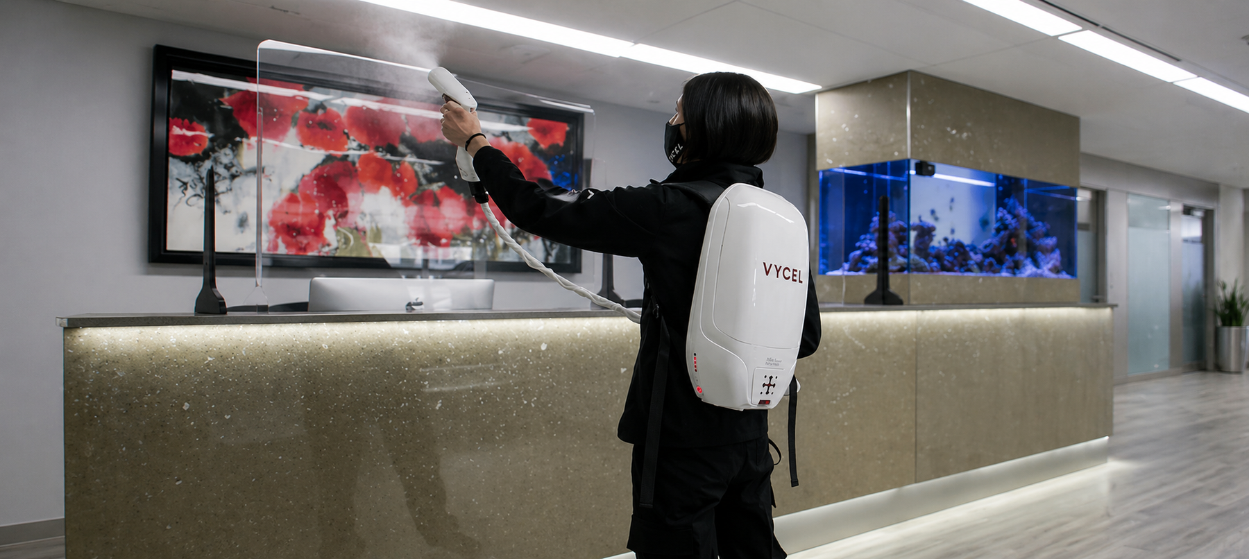 Worker using a Vycel backpack electrostatic sprayer to disinfect a modern reception counter with a protective screen and aquarium in the background.