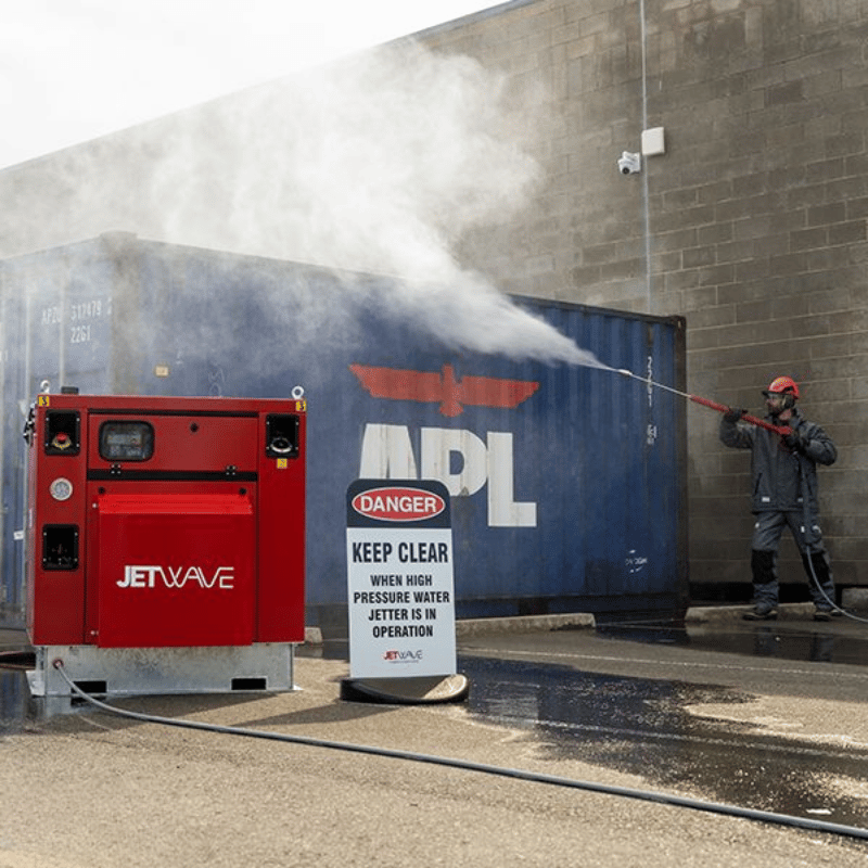 Red Jetwave Executive Silent 280-31 Hot Water Diesel Pressure Cleaner labeled 'JETWAVE' cleaning a large blue container with visible branding, near a 'DANGER' sign.