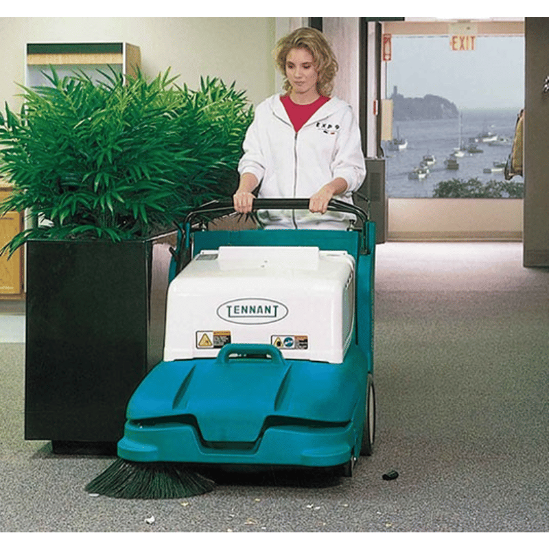 Person operating a Tennant 3640 Walk Behind Floor Sweeper in an indoor setting with a plant and window view.