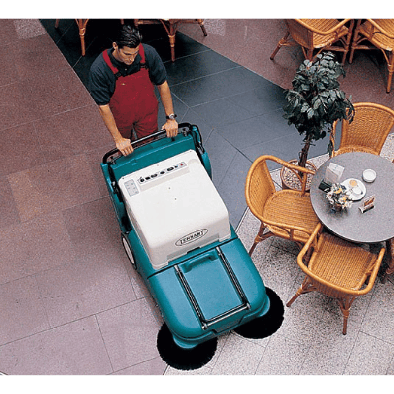 Person using a Tennant 3640 Walk Behind Floor Sweeper in an outdoor setting with tables and chairs.