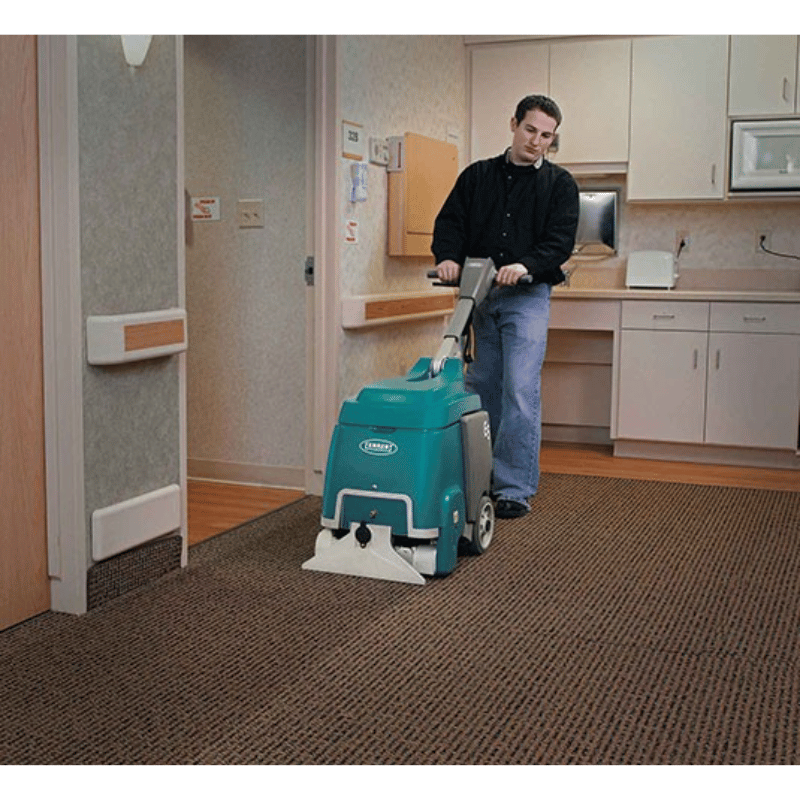 Tennant E5 Compact Walk Behind Carpet Extractor being used by a person in a medical centre on a carpet floor with a kitchen area in the background