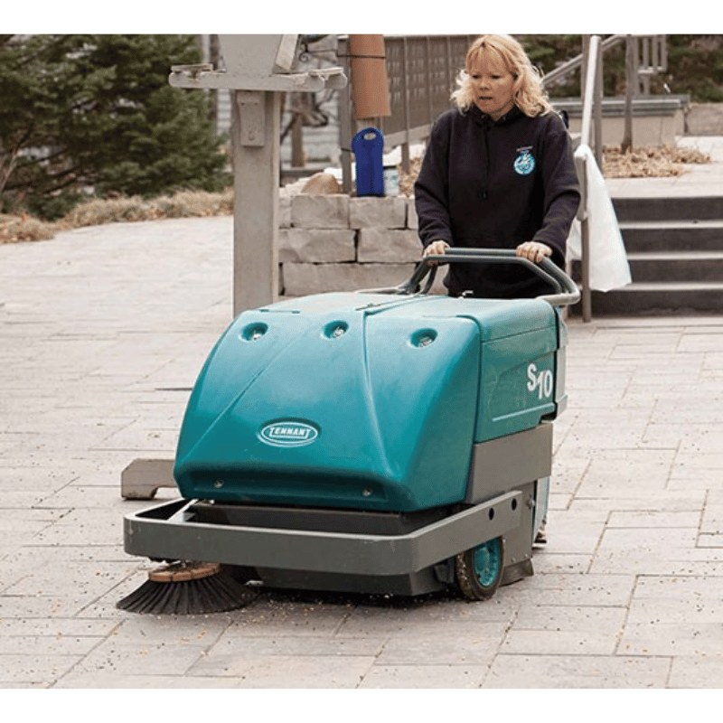 Person pushing a teal and gray Tennant S10 Battery Walk Behind Floor Sweeper on a paved surface.
