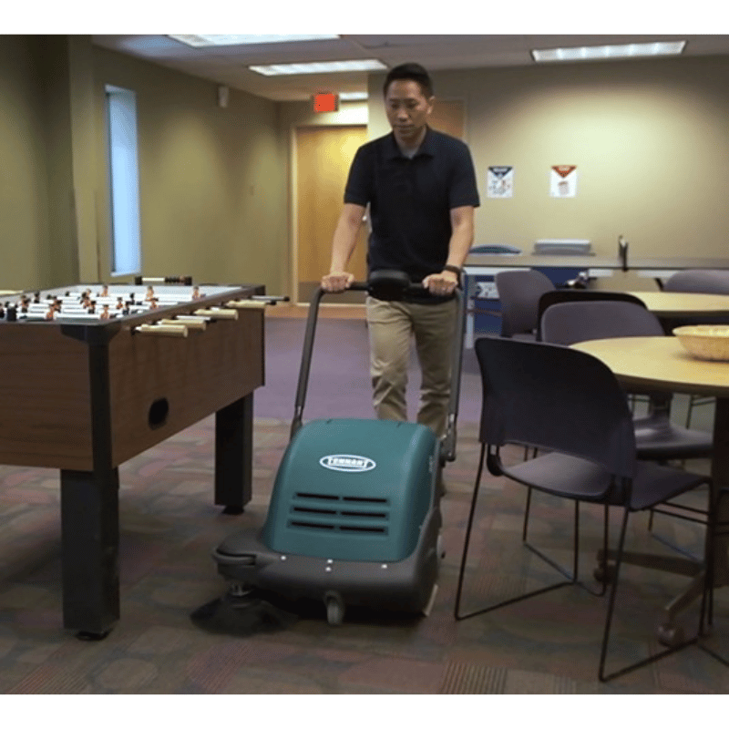 Person operating a Tennant S7 Walk behind floor sweeper in a room with tables and chairs.