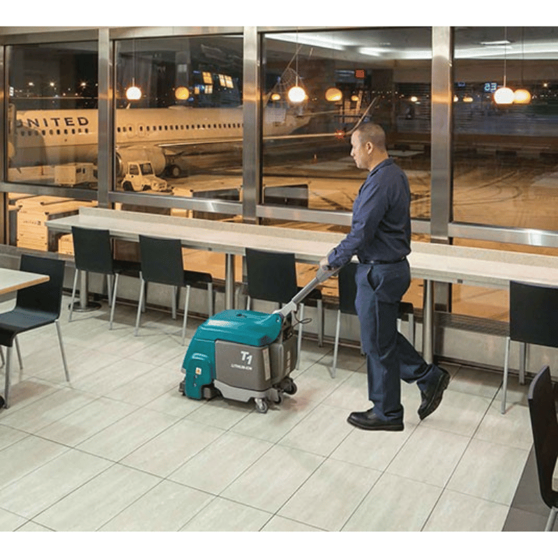 Person cleaning a floor with a Tennant T1B Battery Compact Walk Behind Floor Scrubber in an airport terminal.