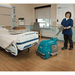 Lady using a Tennant T1B Battery Compact Walk Behind Floor Scrubber to clean the floor next to a hospital bed in a medical facility. 