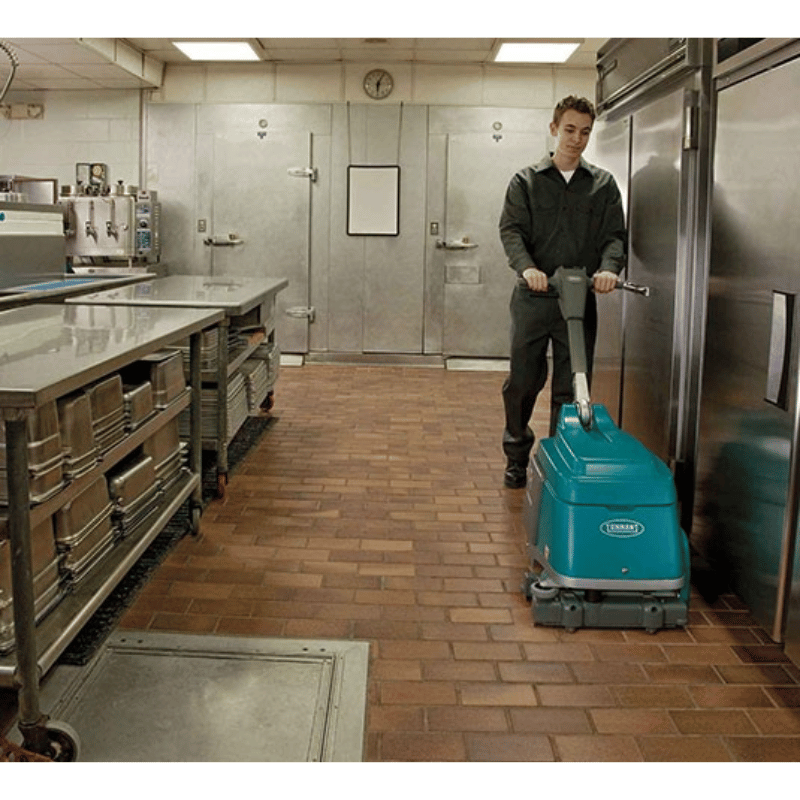 Man using a Tennant T1B Battery Compact Walk Behind Floor Scrubber to clean the floor in a commercial kitchen next to a cold room fridge. 