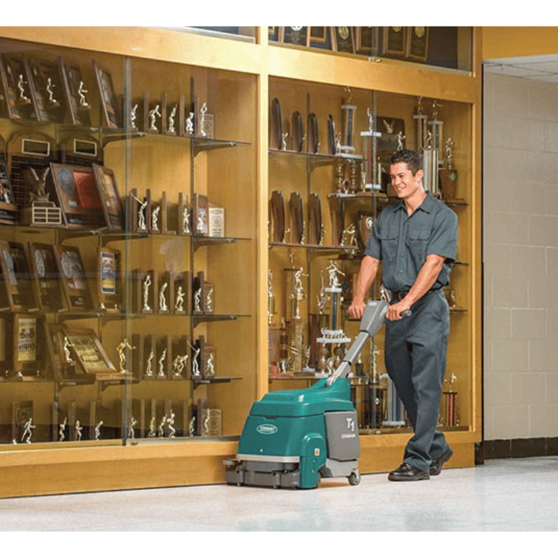 Man using a Tennant T1B Battery Compact Walk Behind Floor Scrubber to clean a hallway next to a trophy cabinet