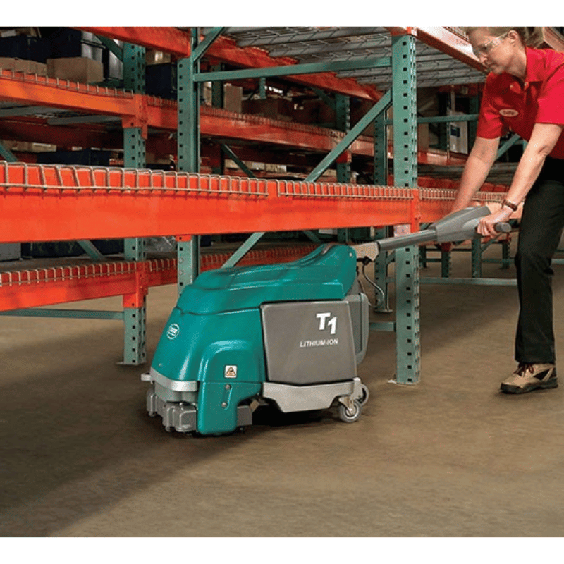 Man using a Tennant T1B Battery Compact Walk Behind Floor Scrubber to clean underneath a shelve in a warehouse facility.
