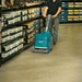 Person using a floor cleaning machine in a warehouse setting with shelves and boxes in the background.