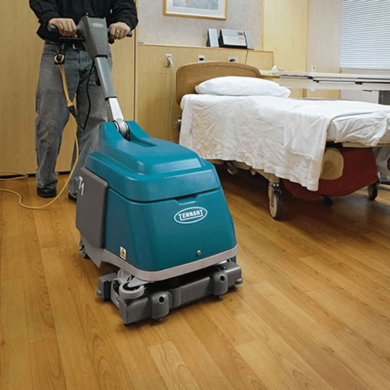 Person operating a teal Tennant floor scrubber in a hospital room with a bed in the background.