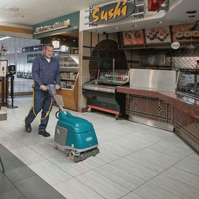 Person using a floor cleaning machine in a shopping centre in front of a sushi restaurant on tiles.