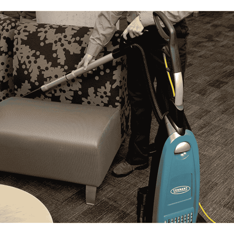 Person using a Tennant VSMU36 Upright Vacuum on a patterned sofa and gray ottoman.